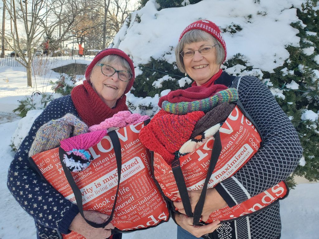 Two women outdoors carrying bags of knit hats