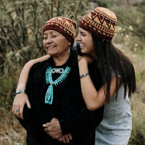 A photo of two Navajo women, one younger and one older. They both wear hand knit hats. 