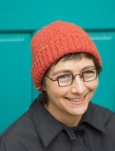 Kate Atherley wearing a black coat, square framed glasses, and an orange knit hat 