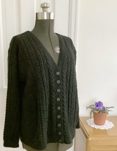 A black cable knit cardigan on a manequin next to a potted plant