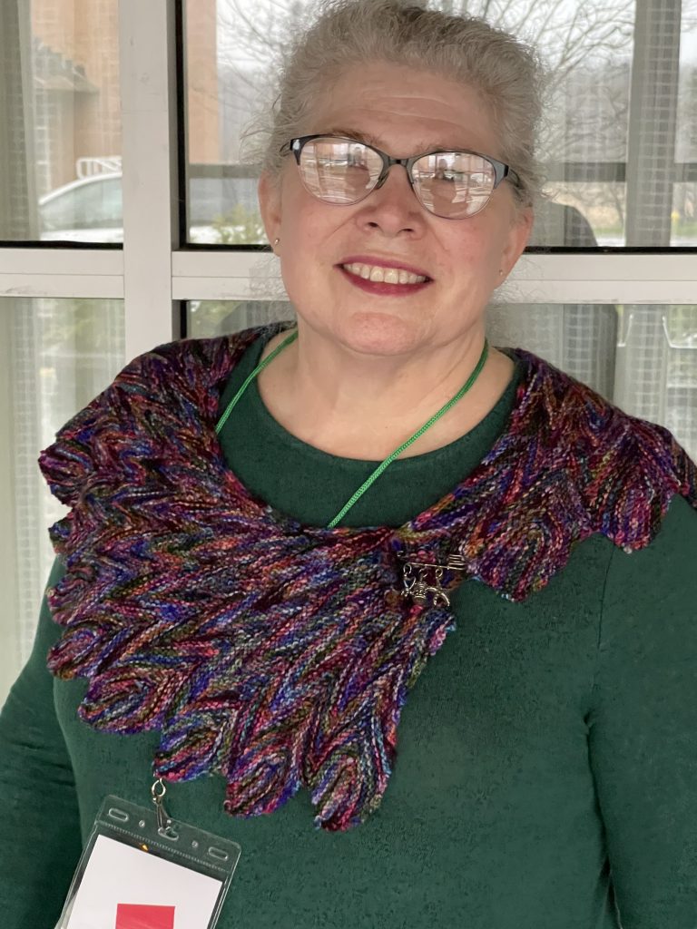 Woman with grey hair and glasses wearing a small knit multi color shawl and green shirt