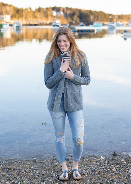 Alicia Plummer standing next to water. She has long blonde hair and wears a hand knit sweater and ripped jeans.