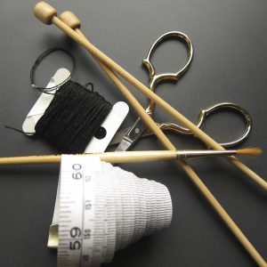 A measuring tape, paint brush, knitting needles, black embroidery thread, and small scissors on a grey background