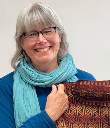 Photo of a woman with grey hair. She wears glasses and a blue scarf. She is holding a brown fair isle sweater
