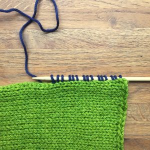 Knitting from green yarn with stitches being picked up along the edge in blue yarn