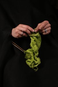 A photograph of hands knitting green yarn against a black background.