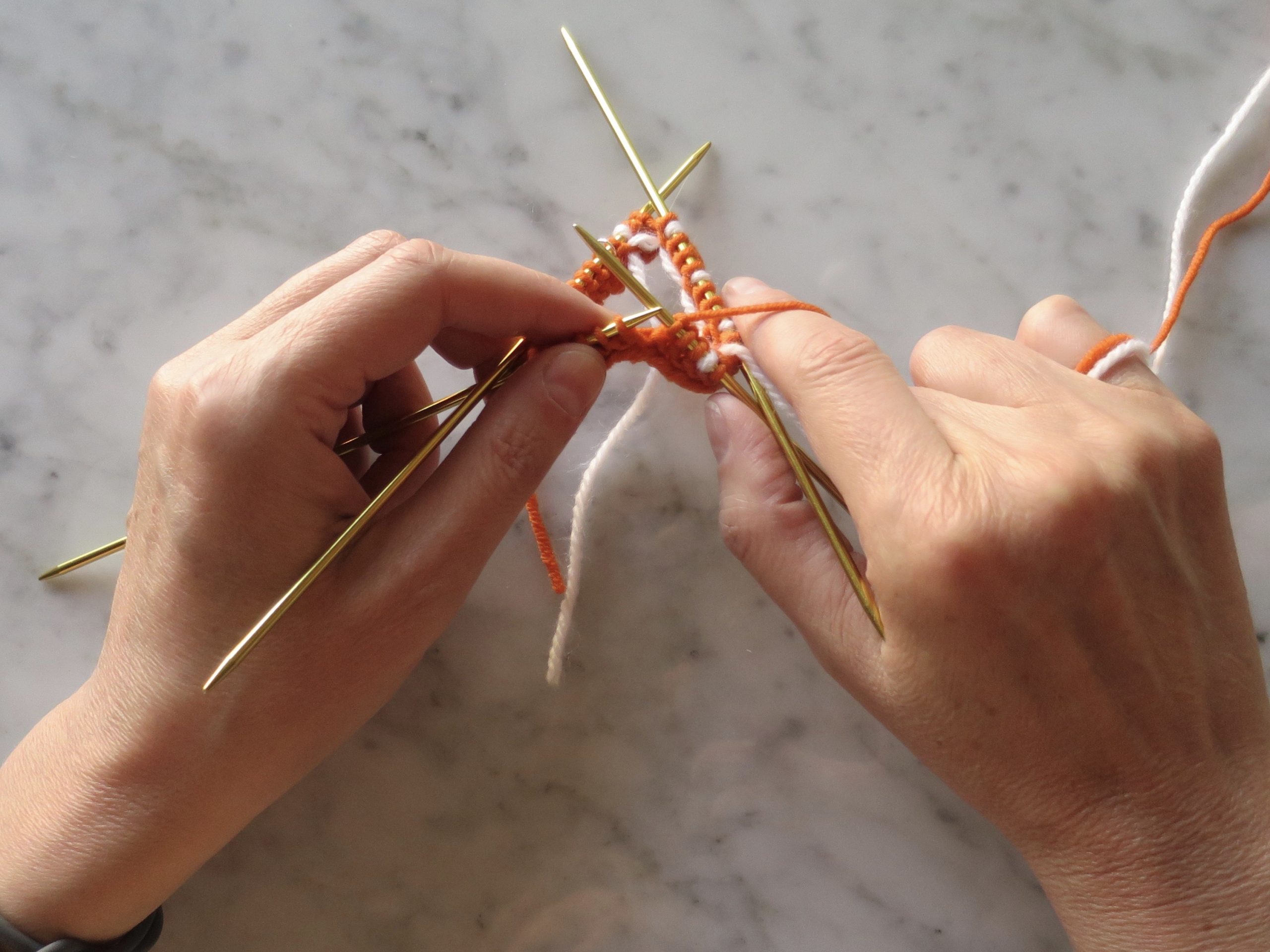 hands knitting in the round on double pointed needles with orange and white yarn
