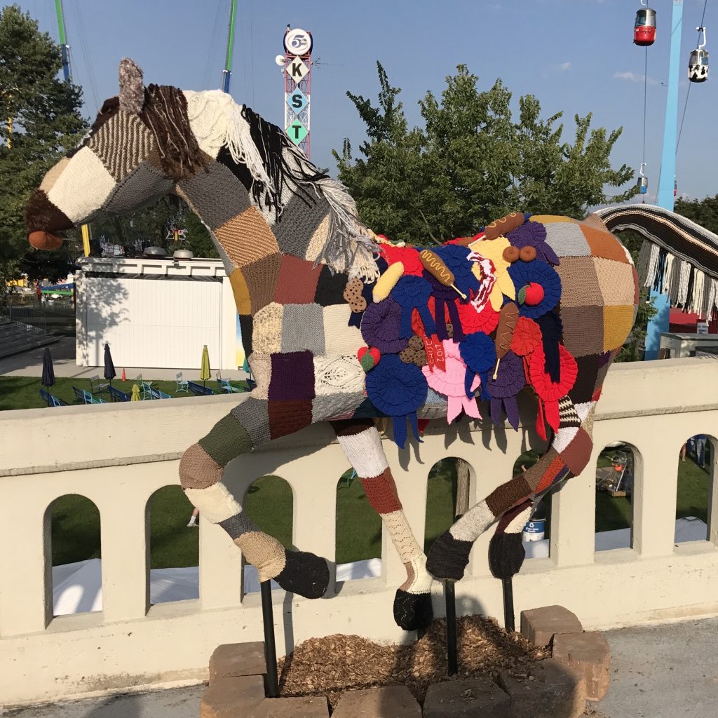 A topiary form of a horse covered in knit squares, ribbons, and fair food. It it on the bridge to the Grandstand building at the fairgrounds.