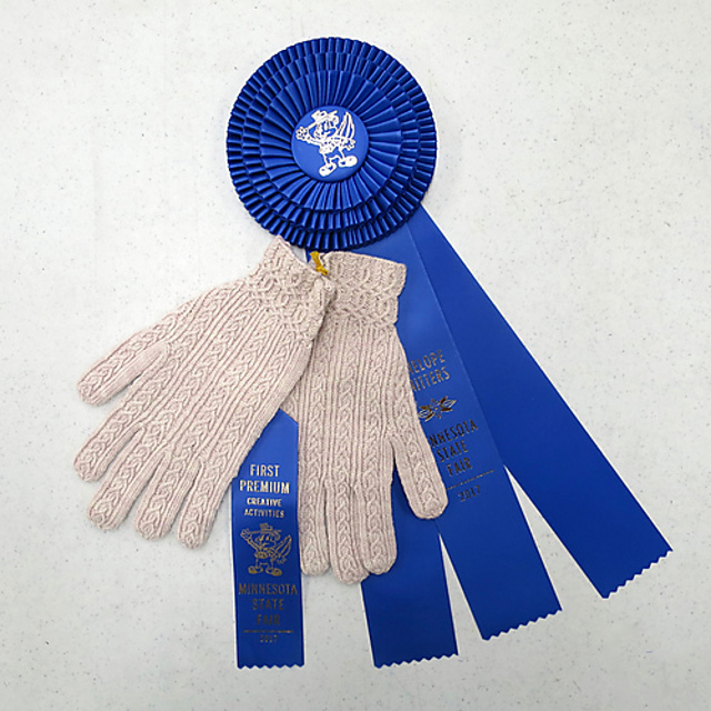 A pair of hand knit white gloves with a state fair ribbon