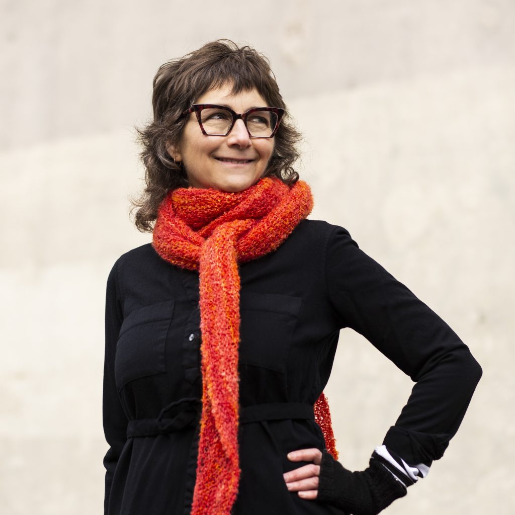 A picture of a woman with shoulder length brown hair. She wears glasses, a black sweater, and an orange scarf.