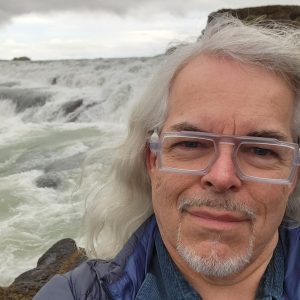 Photo of man with long grey hair in front of rushing water. He wears a blue shirt and clear framed glasses.