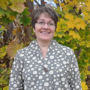 Laurann Gilberston. She has short brown hair and wears glasses. She is standing in front of fall leaves and wears a grey shirt with white polka dots.