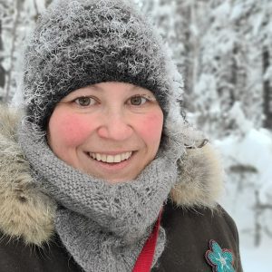 A close up of a woman in a snowy forest wearing a hat, cowl, and parka. Her hat is snow covered