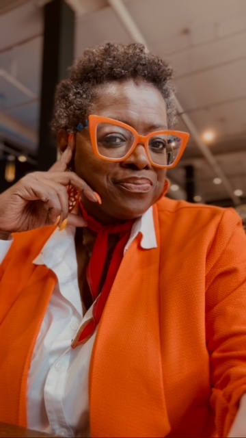 A photo of a woman wearing an orange jacket, white blouse, and orange glasses.